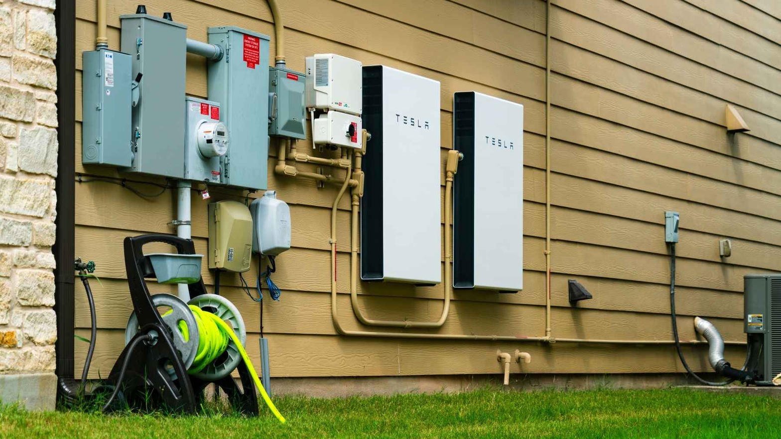 Dwa Tesla Powerwalle na drewnianej ścianie domu, obok skrzynek elektrycznych
